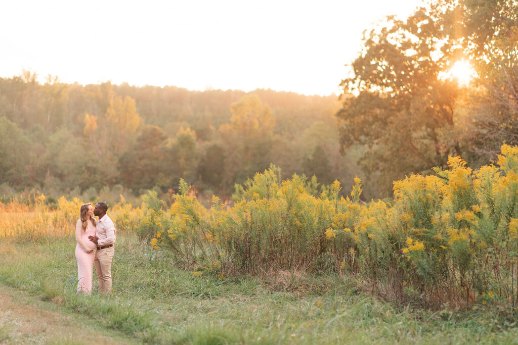 Expecting couple holding hands in a sunlit field during golden hour, mom in flowing pink dress.