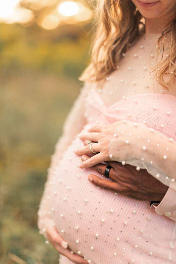Close-up of mom-to-be’s hands cradling her baby bump with soft greenery in background.