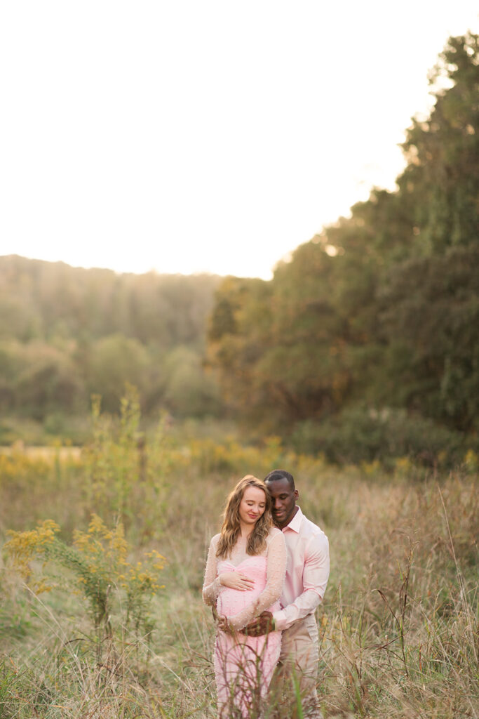 Pregnant woman in pink gown looking down at her belly while partner embraces her from behind
