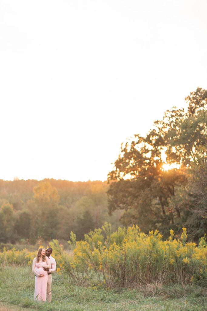Couple smiling at each other with hands on the baby bump, surrounded by nature.
