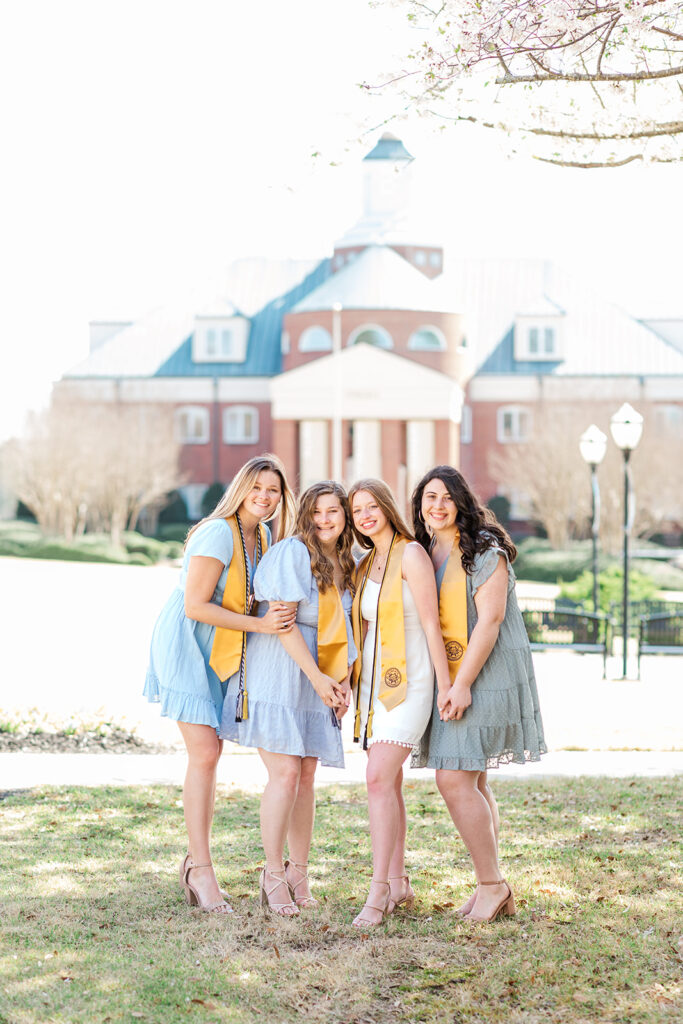 four friends cuddled up together as a group photo with college building behind them during college senior photo shoot in Charlotte, NC