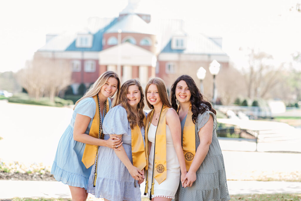 four friends cuddled up together as a group photo with college building behind them during college senior photo shoot in Charlotte, NC