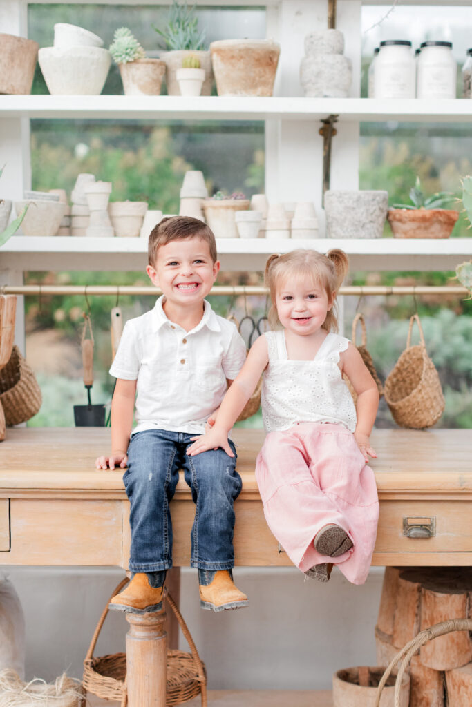 Boy and girl sitting on a side table in Fleurish Greenhouse in Taylorsville, North Carolina both smiling for the camera. 