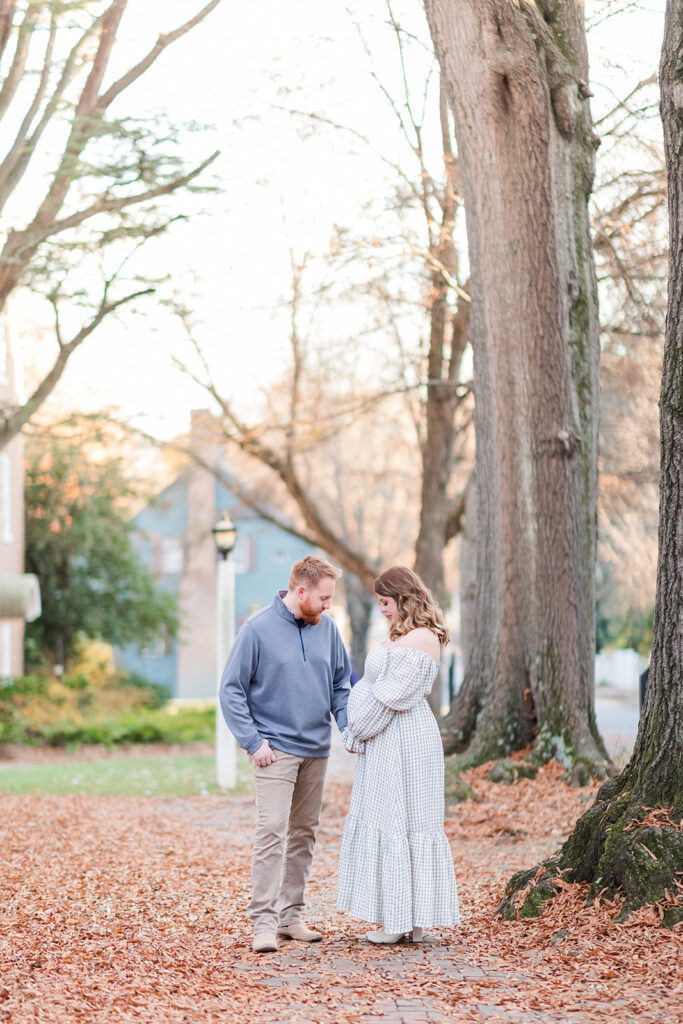 Husband holding hands with wife looking down at her pregnant belly during outdoor maternity photos near Statesville NC.