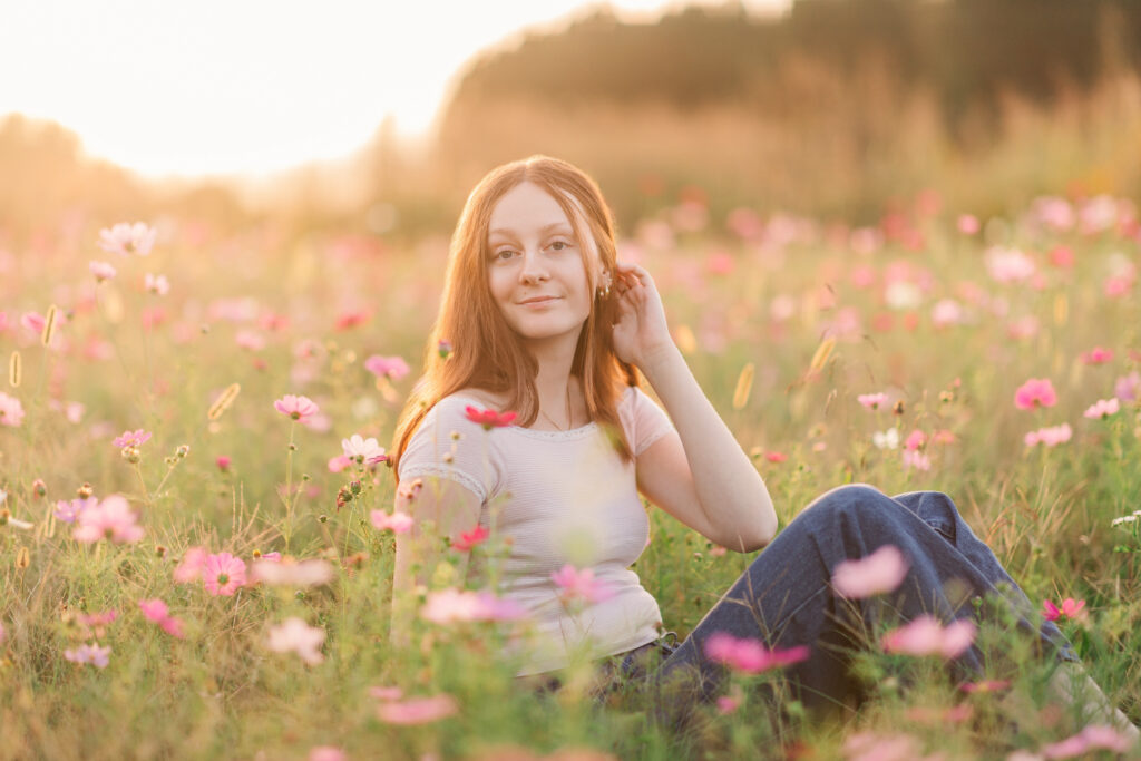 Senior girl sitting in a wildflower field pushing hair back during golden hour wearing a timeless off white shirt and jeans at Windsong Flower Field in Statesville, North Carolina. 
